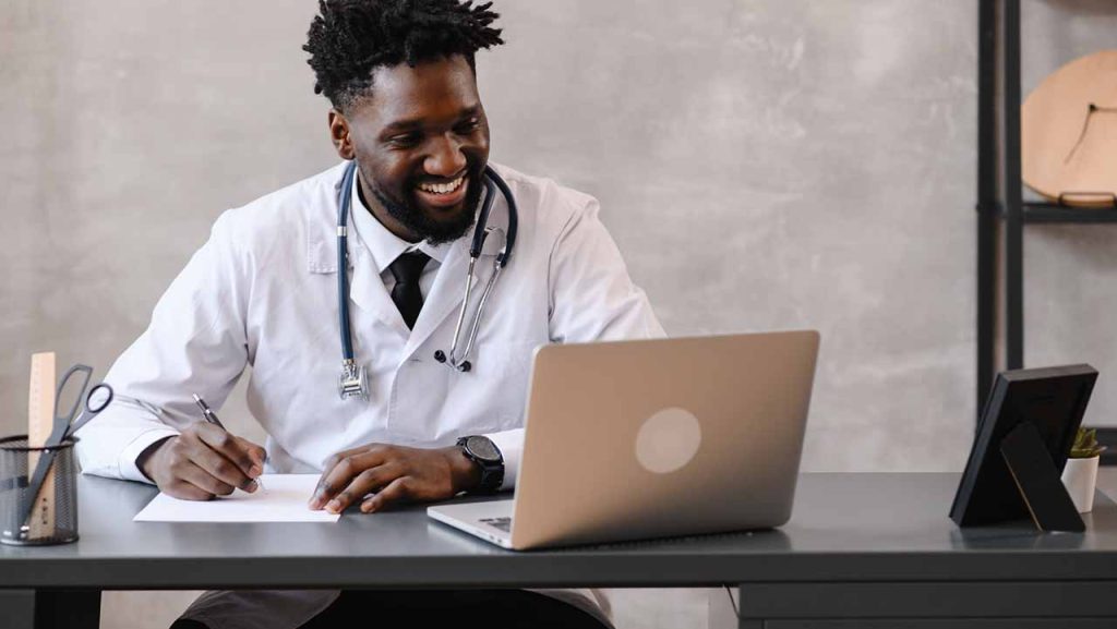 African doctor waering a white coat and stethoscope, sitting at a desk condusting an online consultation